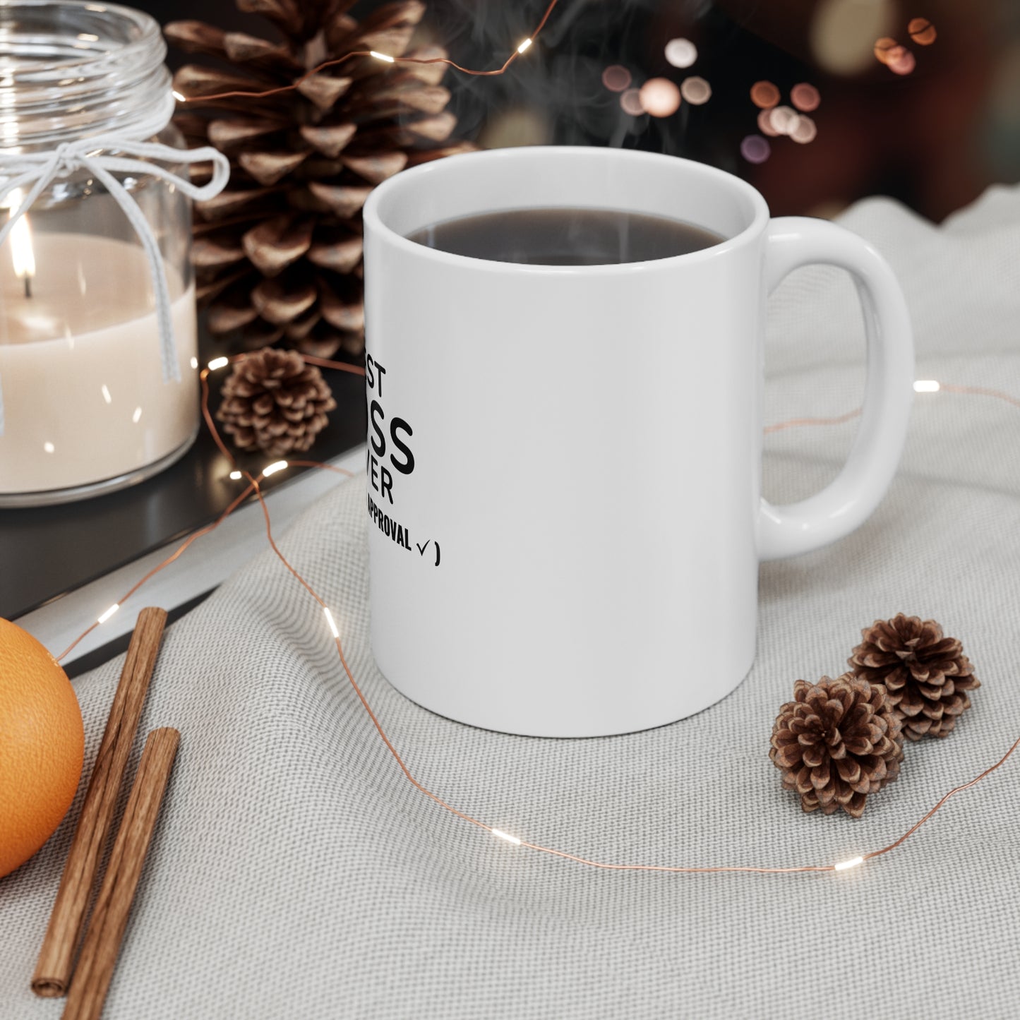White mug with text 'Best Boss Ever (Pending Approval)', candle, cinnamon sticks, and pine cones on a table with string lights.