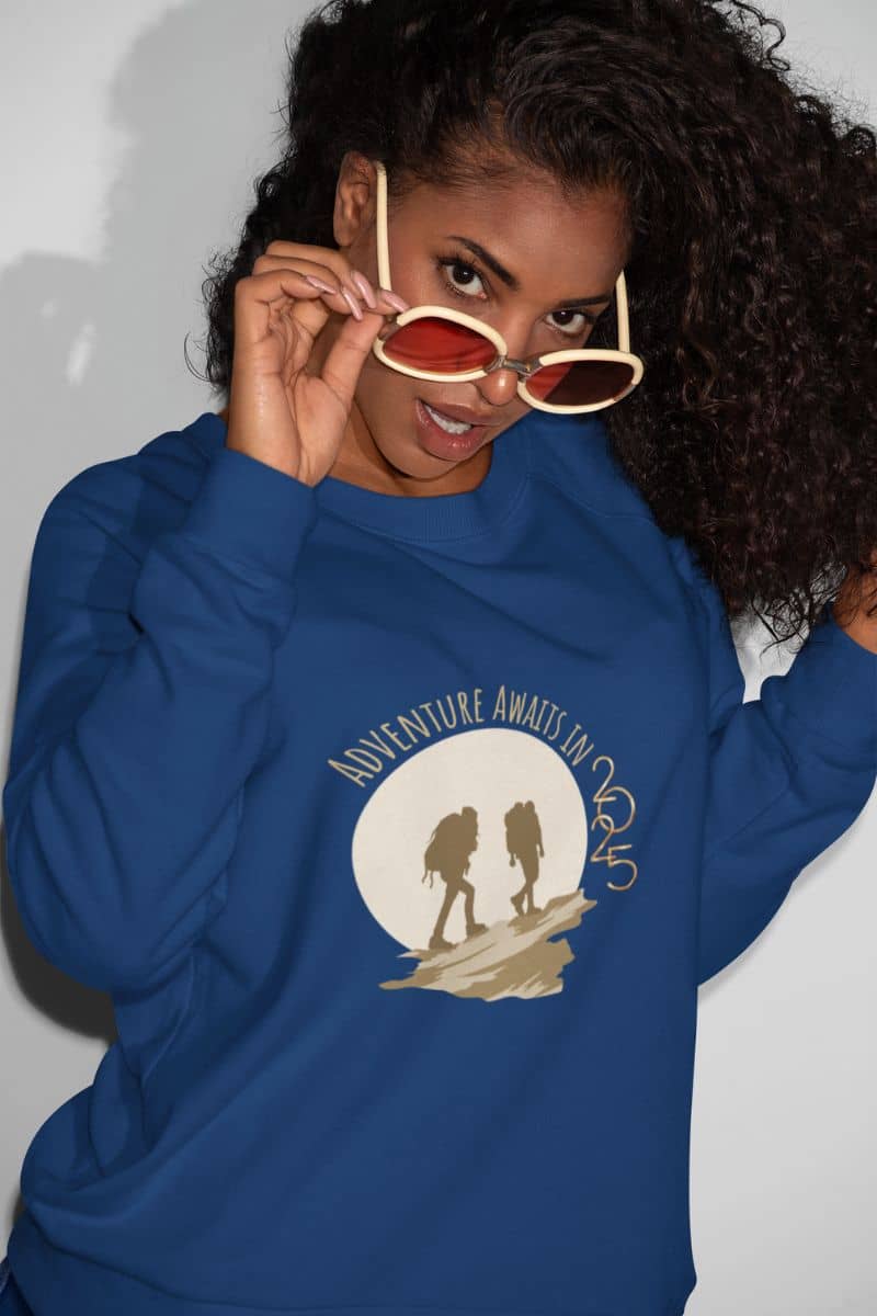 Adventure awaits in 2025 sweatshirt with the girl posing by gently pulling her glasses down her nose