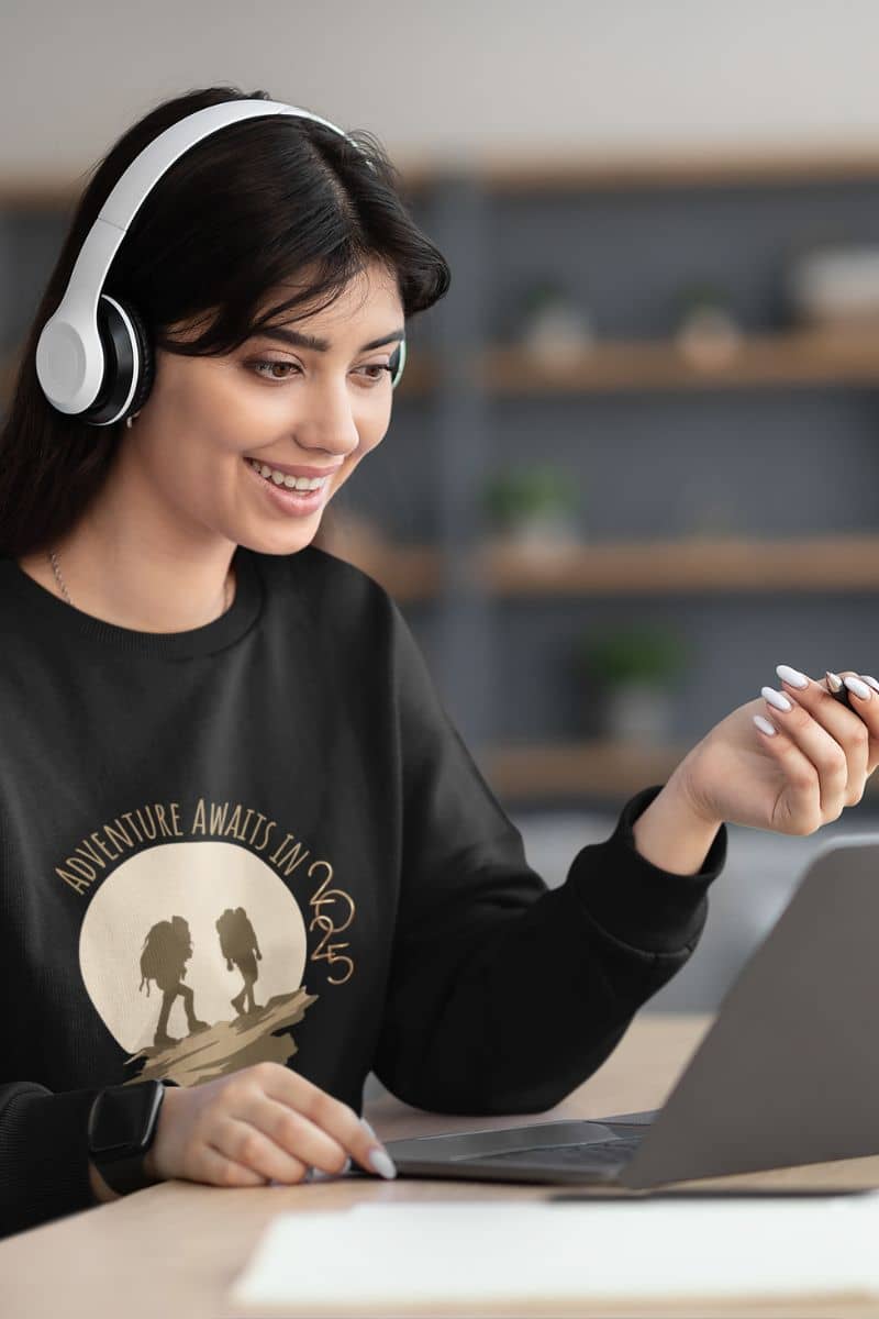 Adventure awaits in 2025 sweatshirt with the girl wearing headphones and looking at the laptop with a bright smile