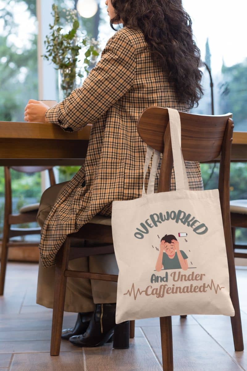 Caffeine addict tote bag with a girl hanging a tote bag on a chair while drinking coffee on the table