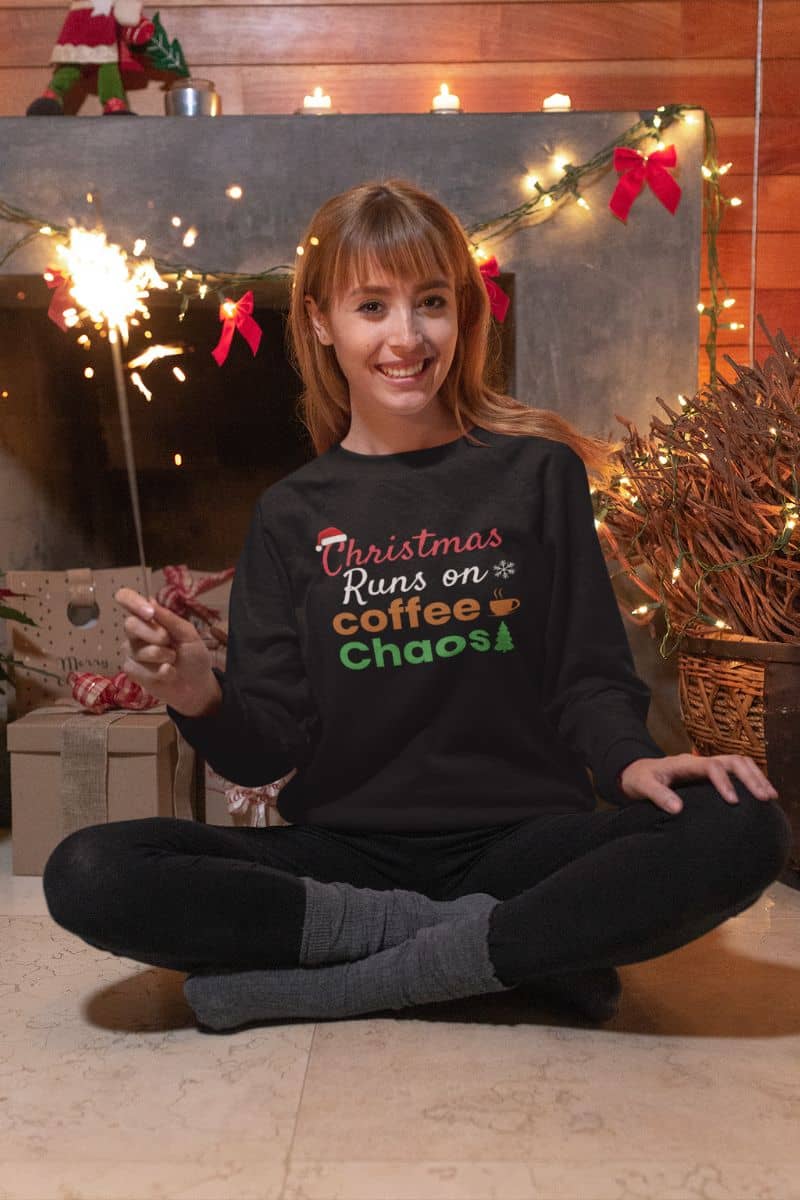 Christmas coffee quote pullover with a woman sitting on the floor wearing a black sweater with text 'Christmas Runs on Coffee & Chaos' in a festive setting.
