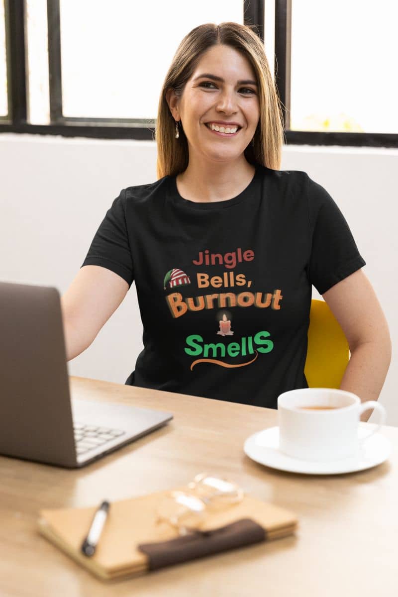 Christmas exhaustion shirt with a person wearing a black t-shirt with humorous text 'Jingle Bells, Burnout Smells', sitting at a desk with a laptop and coffee.