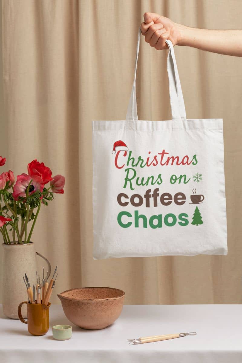Coffee holiday canvas bag with a tote bag with 'Christmas Runs on Coffee Chaos' text held by a hand, with flowers and decor items in the background.