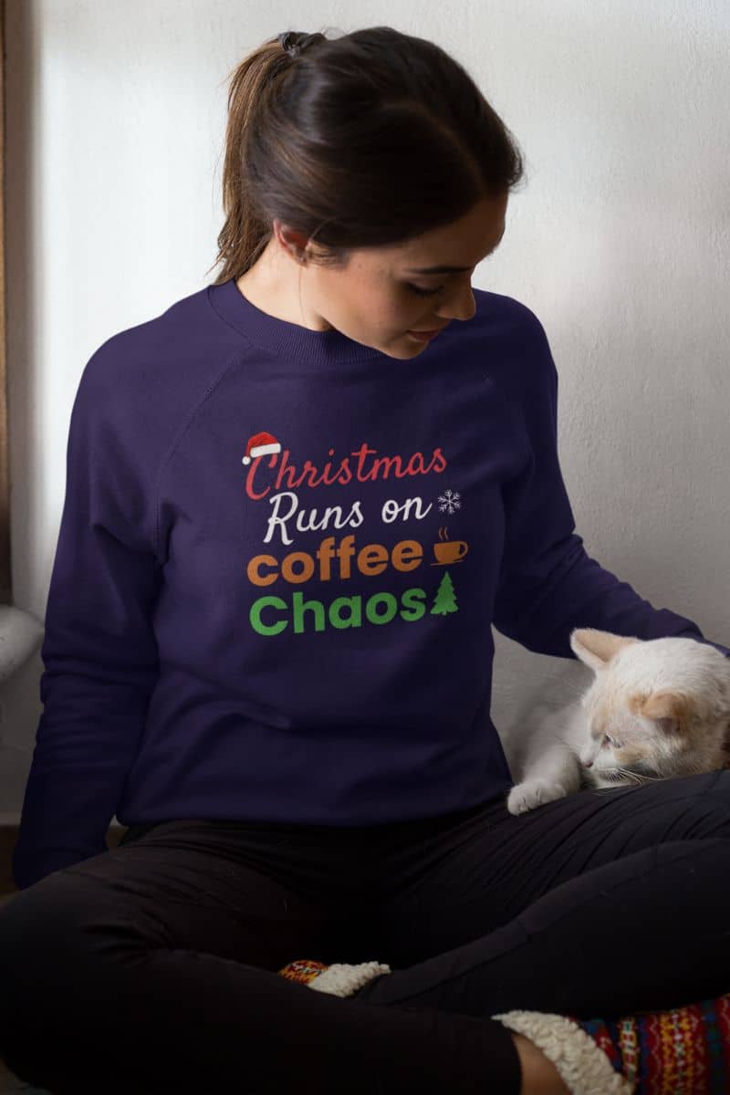 Coffee lover winter sweatshirt with a person wearing a purple sweatshirt with text 'Christmas Runs on Coffee & Chaos' sitting next to a white cat.