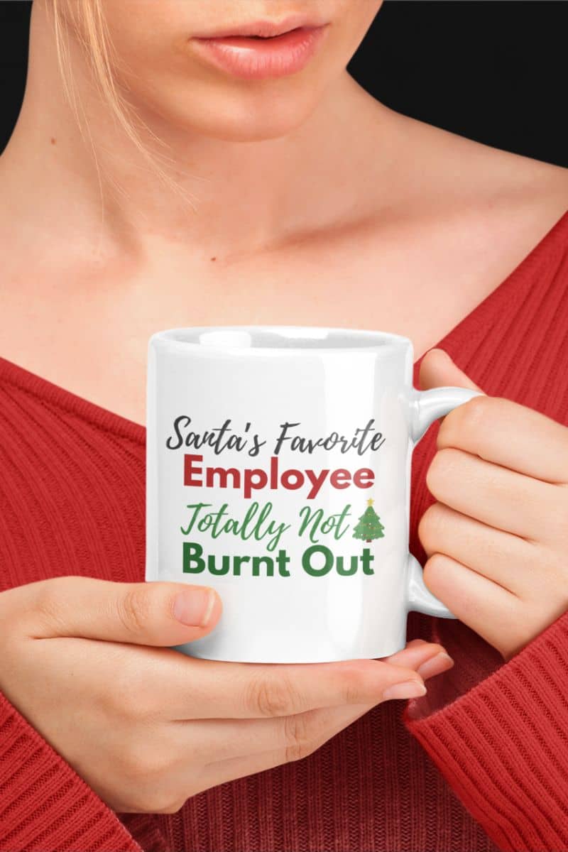 Corporate humor coffee cup with a person holding a white mug with text 'Santa's Favorite Employee Totally Not Burnt Out' in a red sweater.