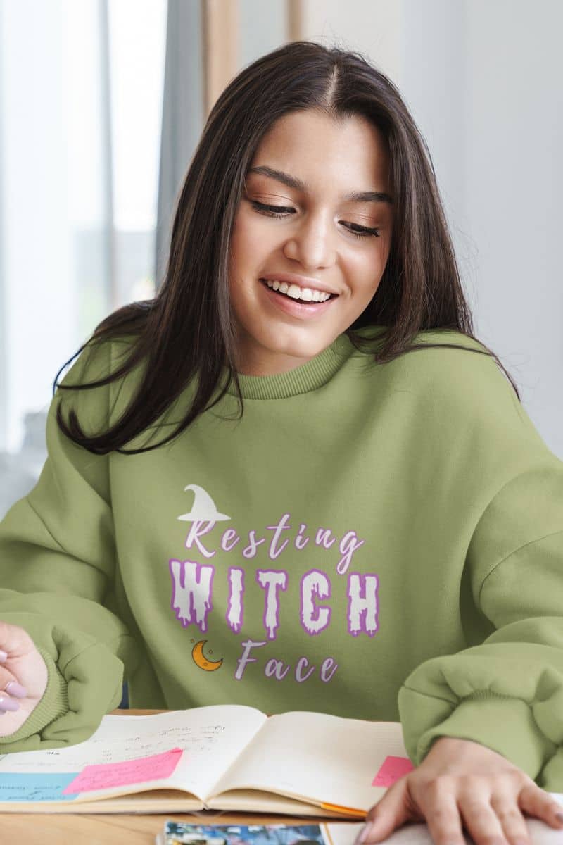 Cozy witch sweatshirt with a girl in a green sweatshirt with 'Resting Witch Face' text, sitting at a table with books.