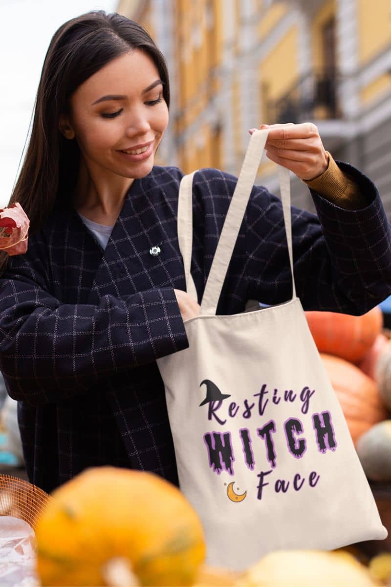 Cute witch shopping bag with a woman holding a tote bag with 'Resting Witch Face' text amidst pumpkins and autumn decor.