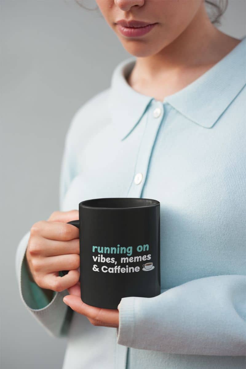 Daily survival mug with a person holding a black mug with text 'running on vibes, memes & caffeine' against a neutral background