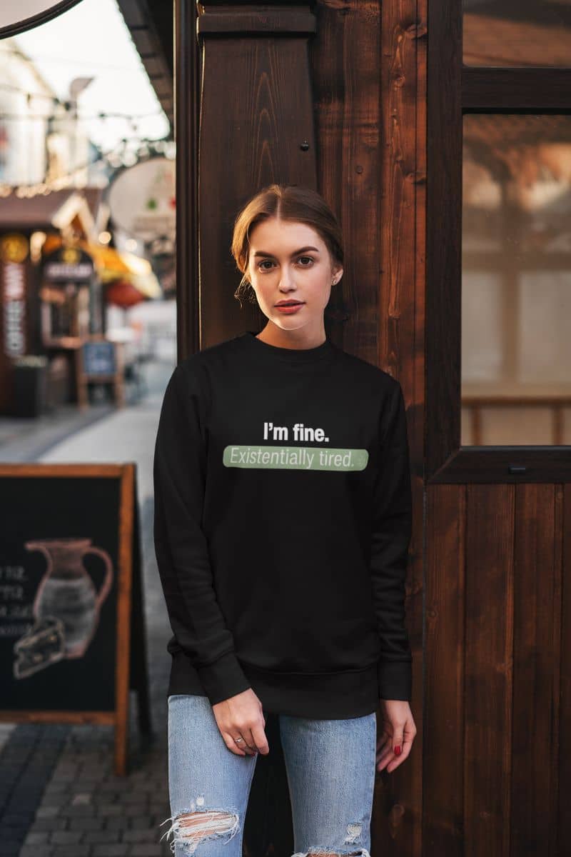 Existentially tired sweatshirt with a person wearing a black sweatshirt with text 'I’m fine. Just existentially tired', standing in front of a wooden door.