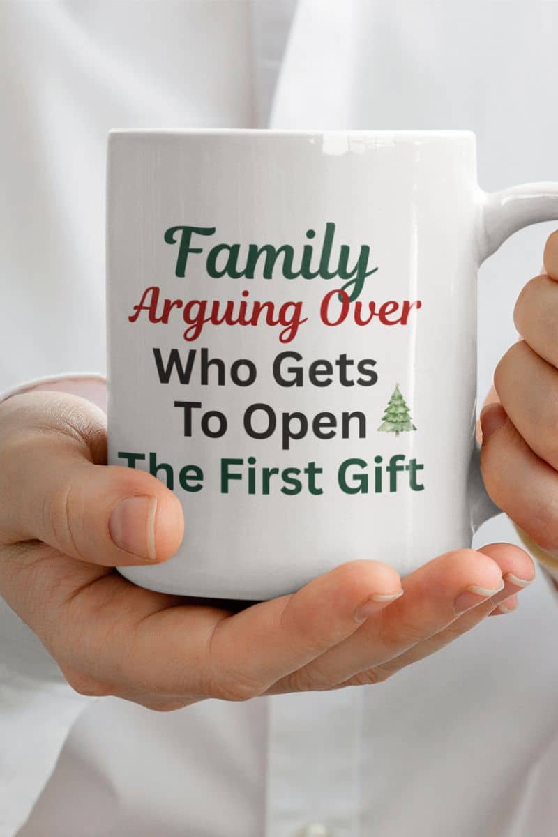 Funny Christmas mug for family with a white mug held by hands with text 'Family: Arguing Over Who Gets To Open The First Gift'