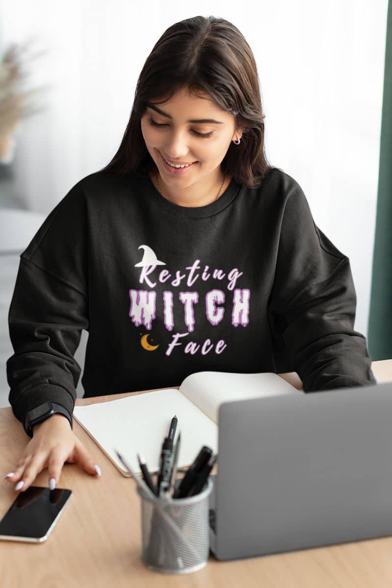Funny Halloween hoodie with a woman wearing a black sweatshirt with 'Resting Witch Face' text, sitting at a desk.