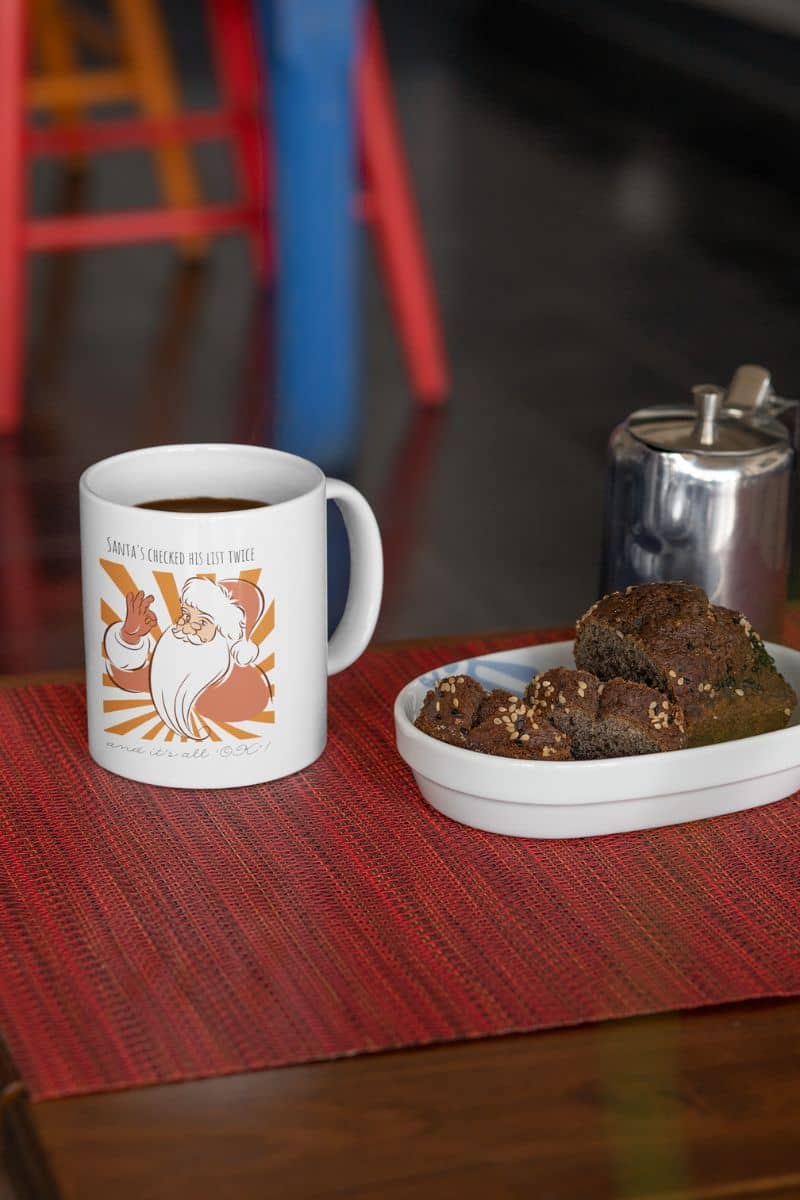 Funny Santa Mug - Santa's Checked His List Twice And It's All OK with a cup of coffee and a plate of cake on the table for breakfast
