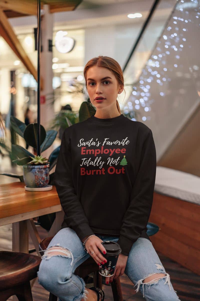 Funny burnout sweatshirt with a woman wearing a black sweatshirt with text 'Santa’s Favorite Employee Totally Not Burnt Out', sitting in a casual indoor setting.