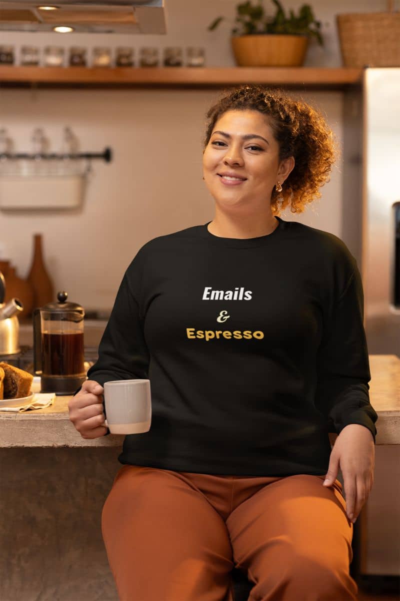 Funny caffeine hoodie with woman sitting in a kitchen holding a coffee cup, wearing a black sweatshirt with 'Emails & Espresso' text.