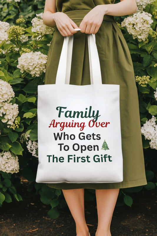 Funny family christmas tote bag with a person holding a tote bag with humorous text 'Family: Arguing Over Who Gets To Open The First Gift' in front of flowers