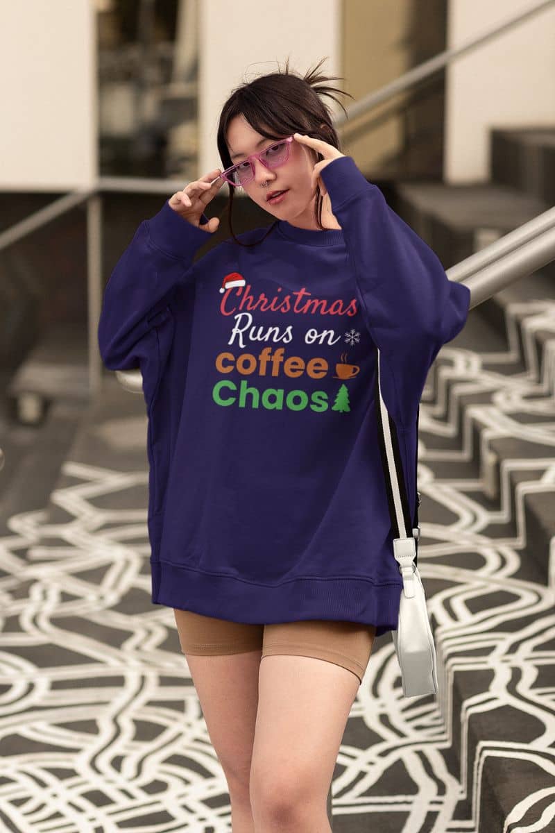Funny holiday sweater with a person wearing a purple sweatshirt with text 'Christmas Runs on Coffee & Chaos', standing on a patterned floor.
