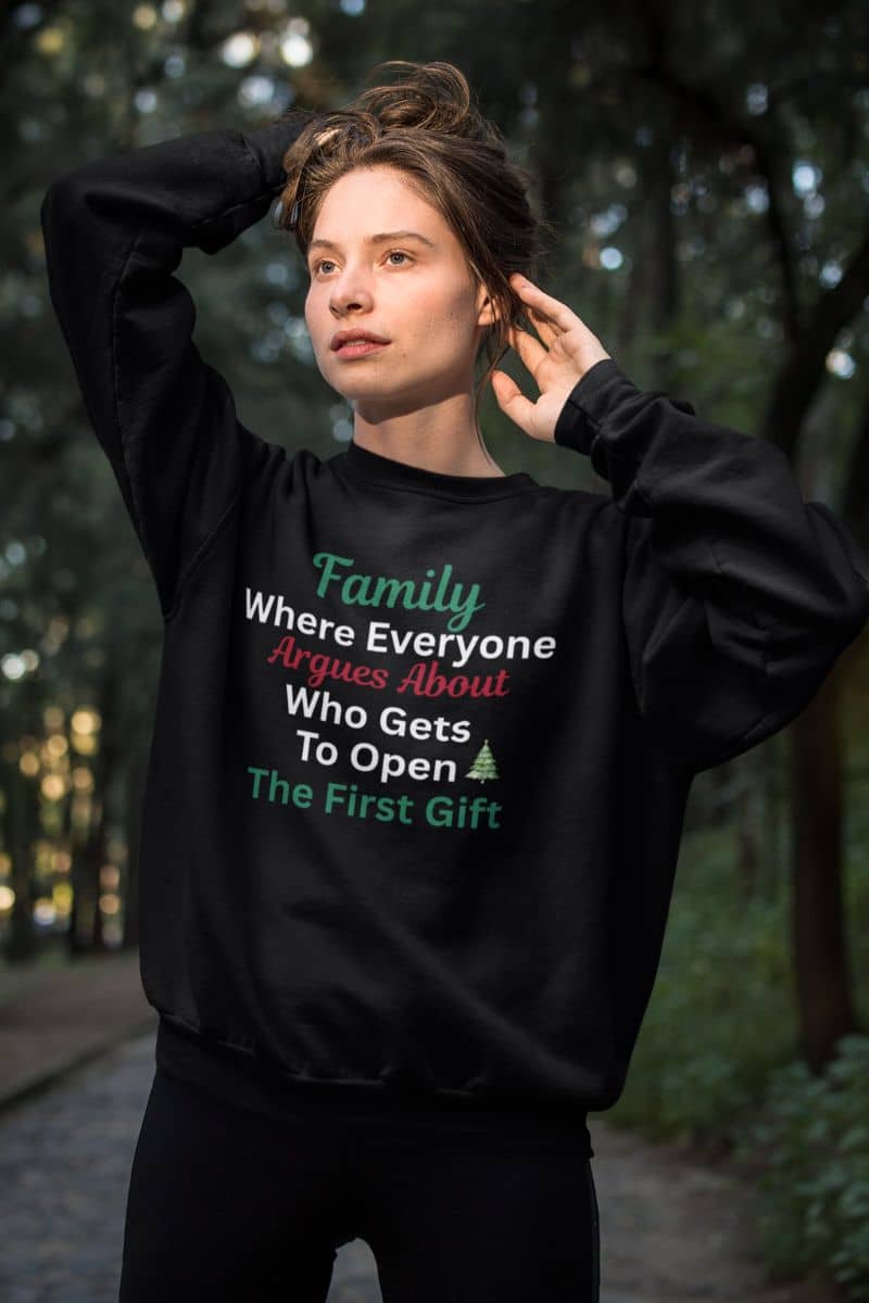 Funny holiday sweatshirt with a person wearing a black sweatshirt with humorous text 'Family: Where Everyone Argues About Who Gets To Open The First Gift' in a forest setting