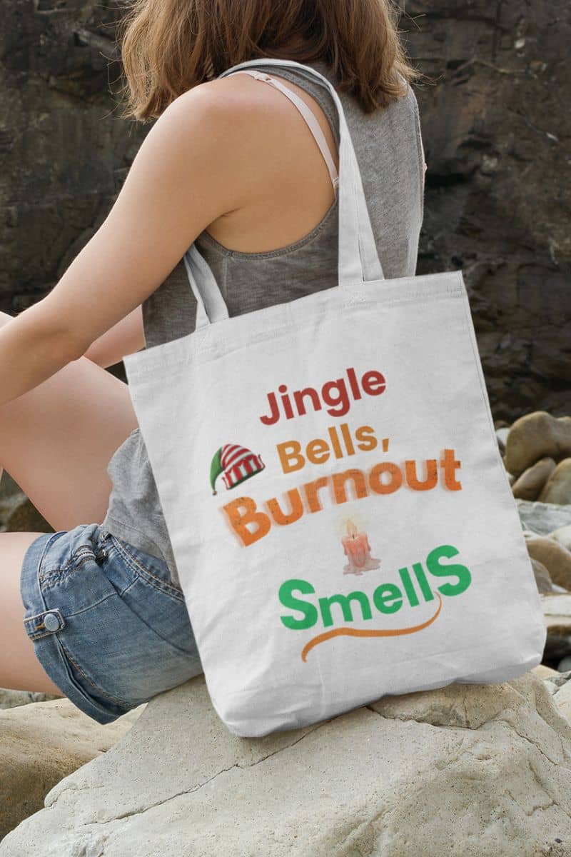 Funny work stress tote bag with a person holding a tote bag with humorous text 'Jingle Bells, Burnout Smells' on a rocky surface
