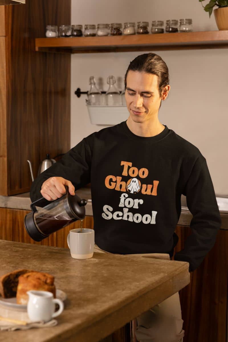 Ghost graphic sweater with a person wearing a black sweatshirt with 'Too Ghoul for School' text, pouring coffee in a kitchen.