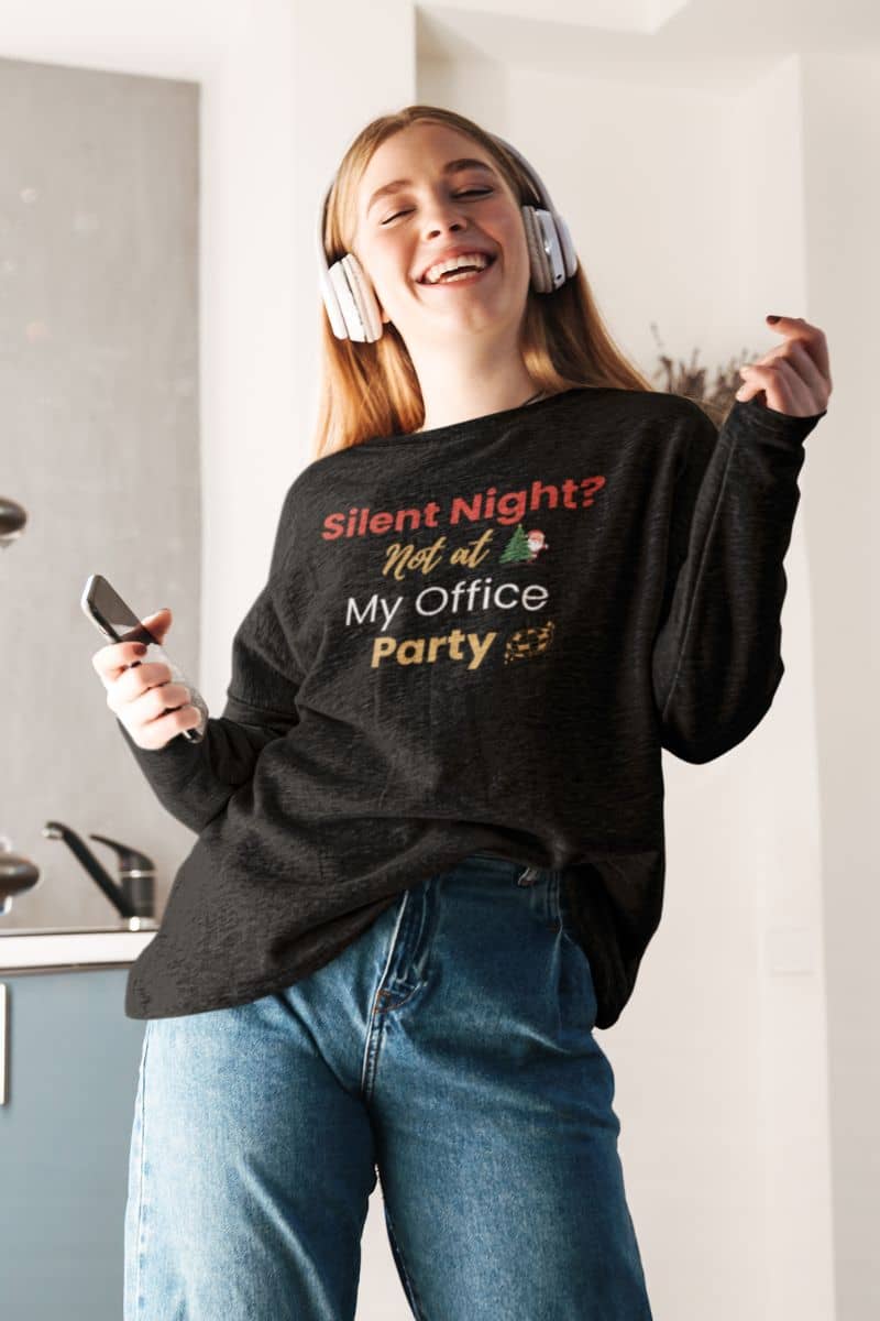 Holiday sweatshirt with a person wearing a black sweatshirt with humorous text 'Silent Night? Not at My Office Party', listening to music in a kitchen.