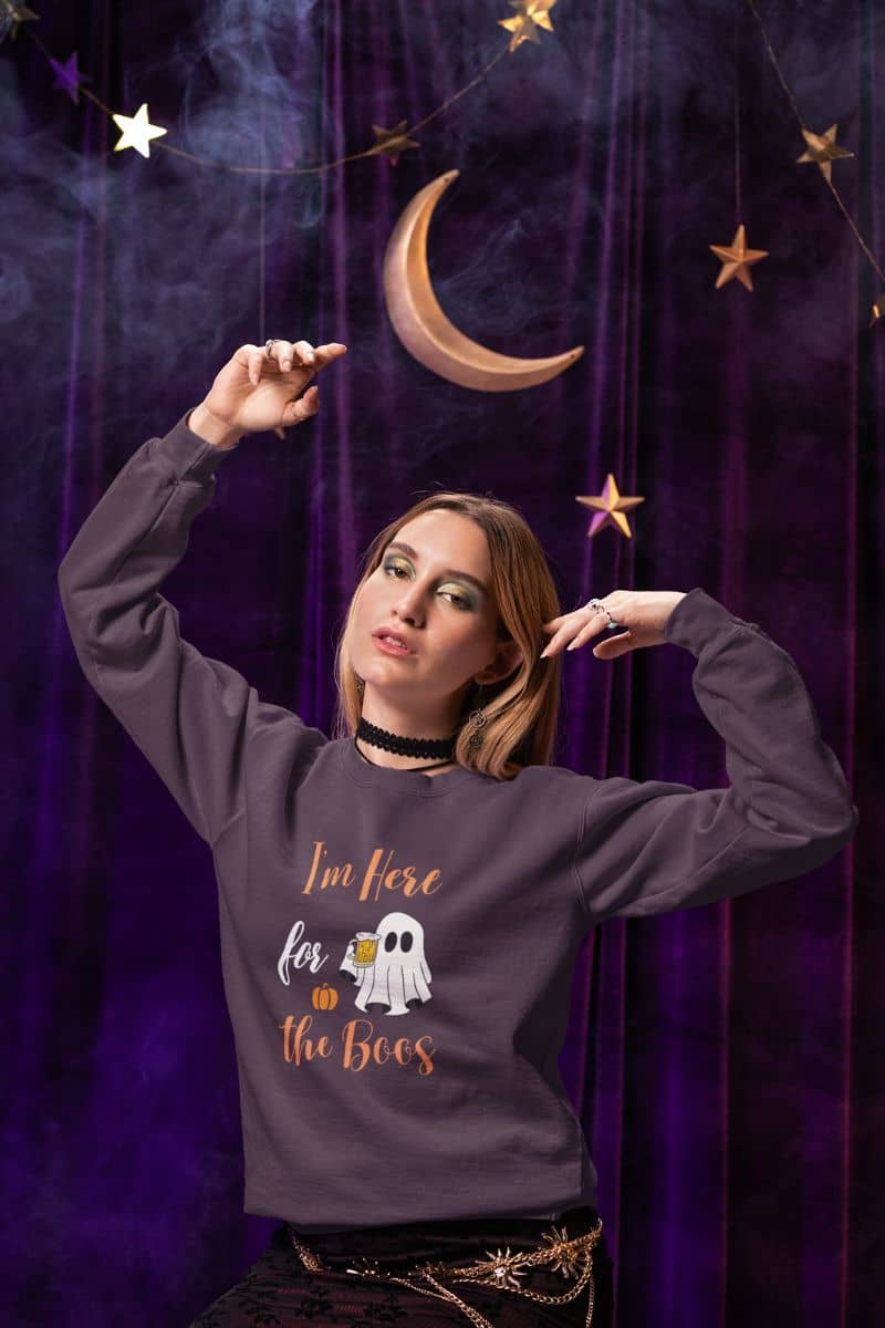 I'm Here for the Boos sweatshirt with a woman wearing a Halloween-themed sweatshirt with a purple curtain and decorative moon and stars in the background.