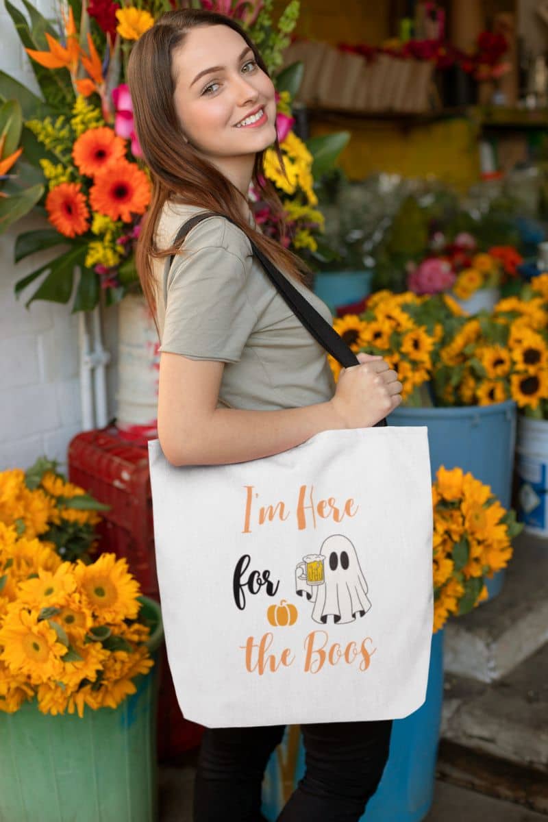 I'm Here for the Boos tote with a woman holding a tote bag with Halloween text in a floral setting