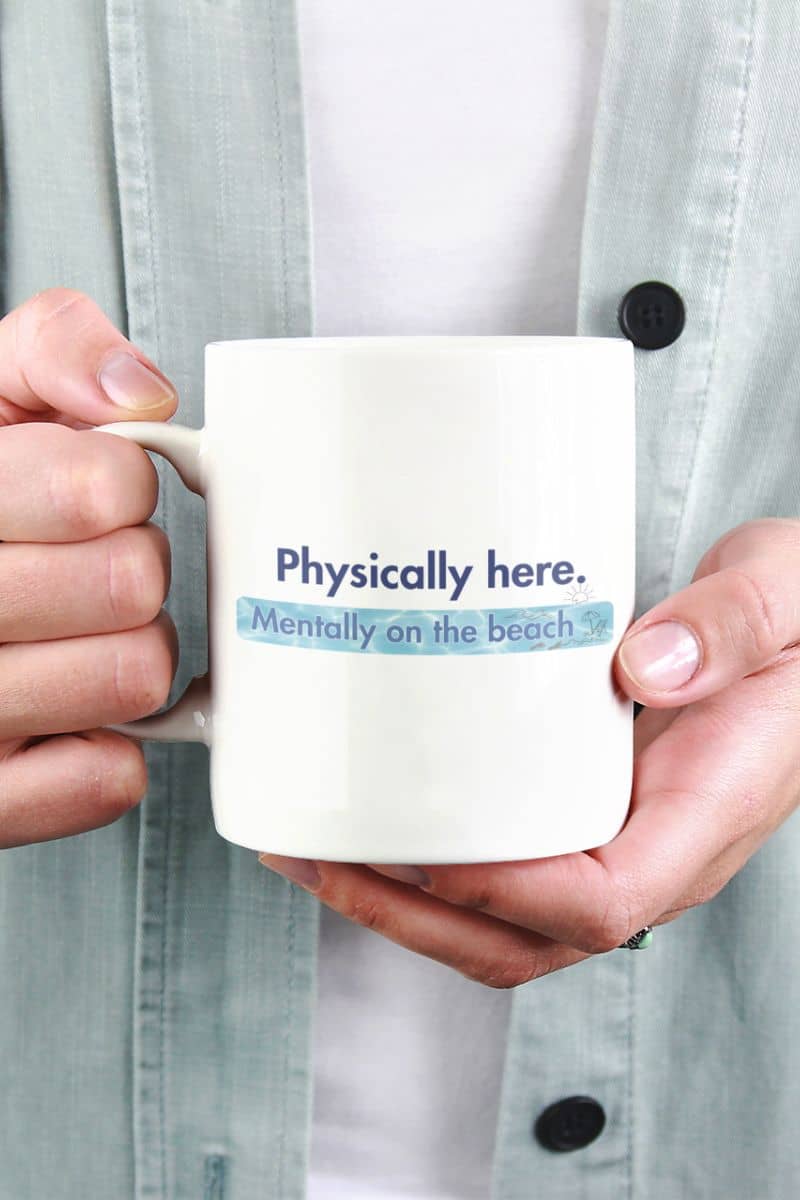 "Mentally on the Beach" coffee mug, with a person holding it and showing the text "Physically here. Mentally on the beach.