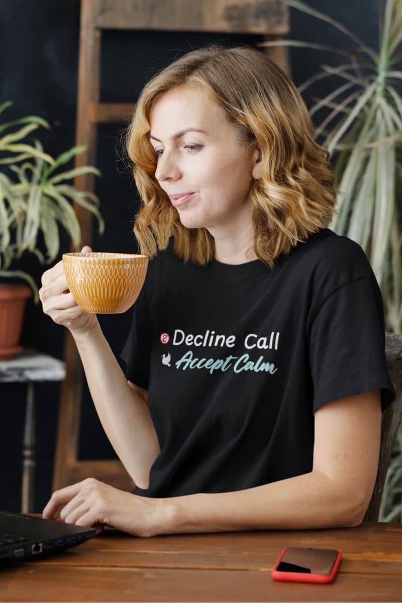Mindful lifestyle tee with a woman holding a mug with text 'Decline Call Accept Calm' on her shirt, sitting at a table with plants in the background