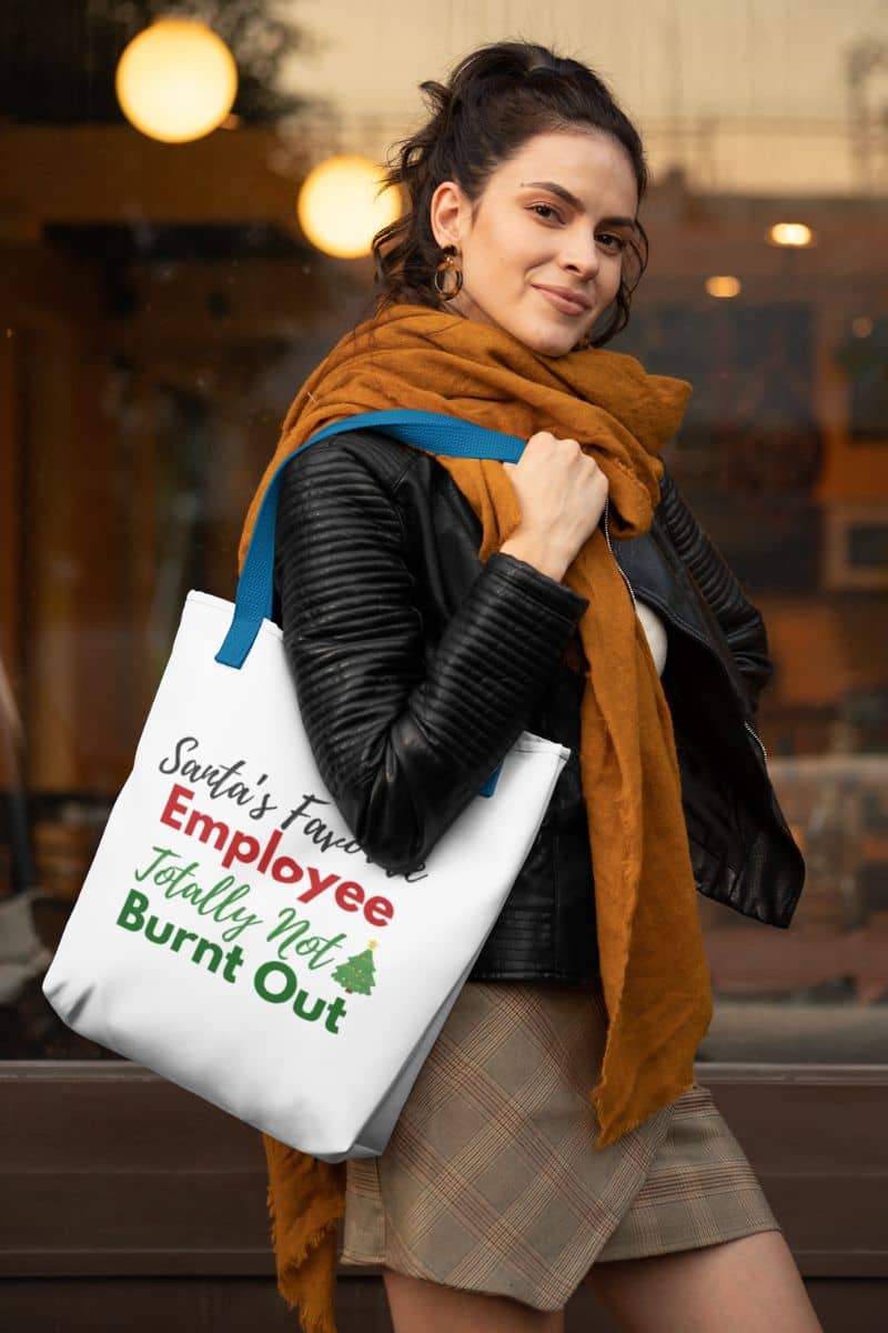 Minimalist office tote showing a woman holding a tote bag with text 'Santa’s Favorite Employee Totally Not Burnt Out', wearing a scarf and jacket, in an indoor setting.