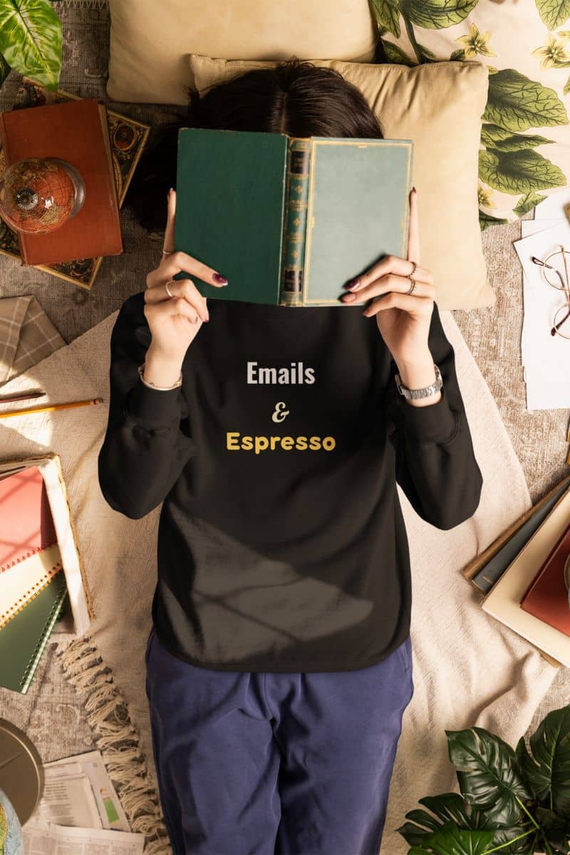 Morning fuel sweatshirt with a person wearing a black shirt with 'Emails & Espresso' text, holding a book over their face in a cozy room.