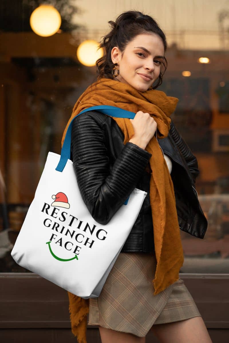Neutral Holiday bag with a woman holding a tote bag with 'Resting Grinch Face' text and Santa hat graphic.