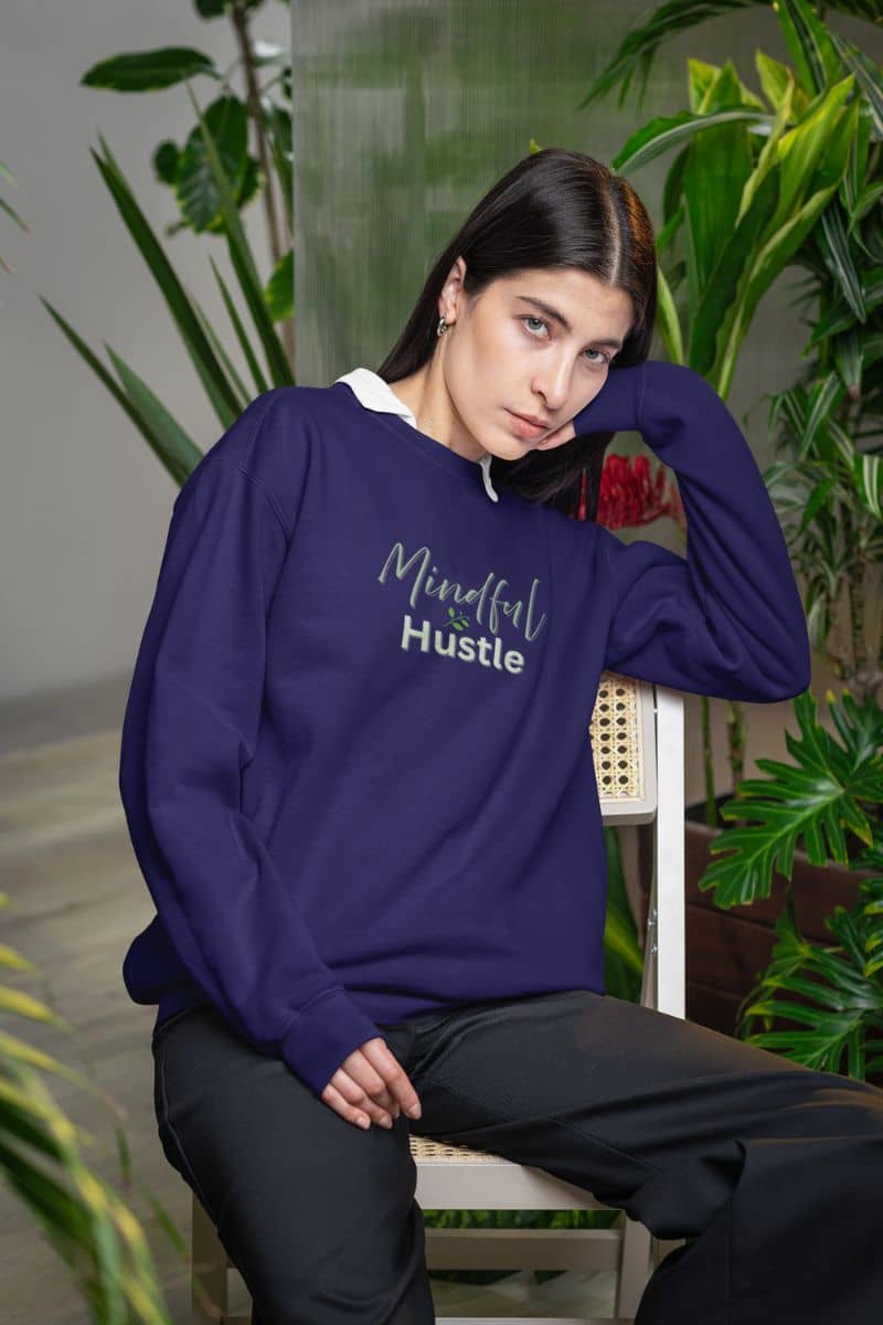 Personal growth hoodie with a person wearing a purple sweatshirt with 'Mindful Hustle' text, sitting among plants.