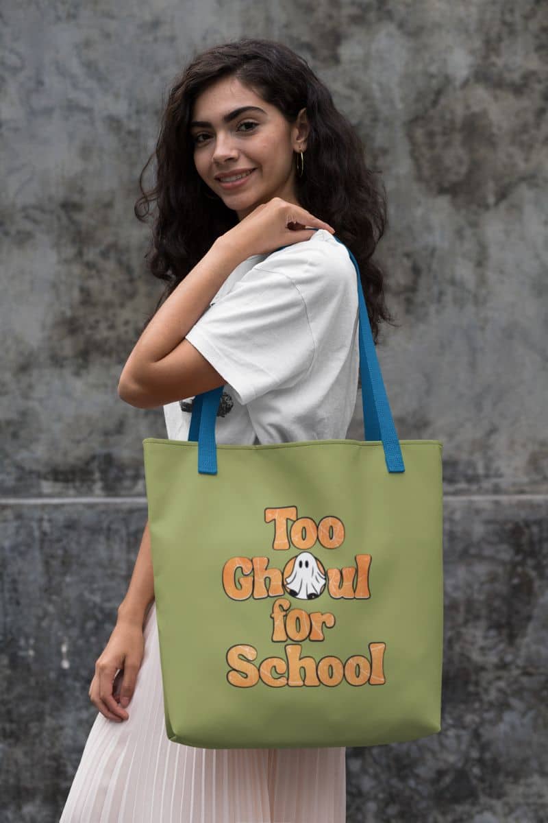 Pumpkin tote bag with a woman holding a green tote bag with 'Too Ghoul for School' text against a textured wall.