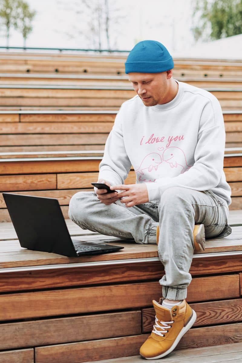 Romantic Dolphin love sweatshirt with a man looking at his phone next to a laptop on a wooden bench