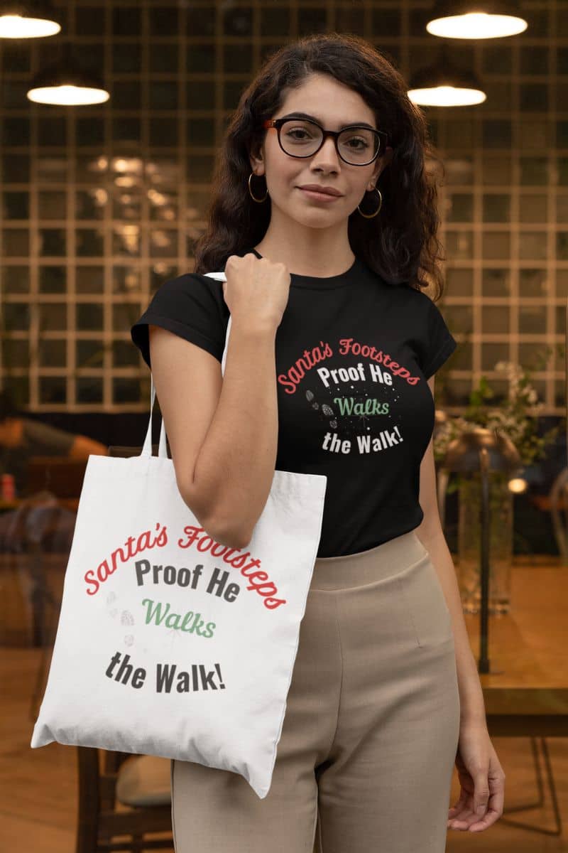 Santa walk tote with a woman wearing a black t-shirt and holding a tote bag with text 'Santa's Footsteps - Proof He Walks The Walk!', standing indoors.