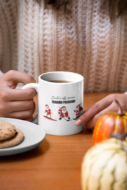 Santa's Off-Season Training Coffee Mug 11oz with a girl holding a cup of coffee on a table with cookies and pumpkins during halloween season