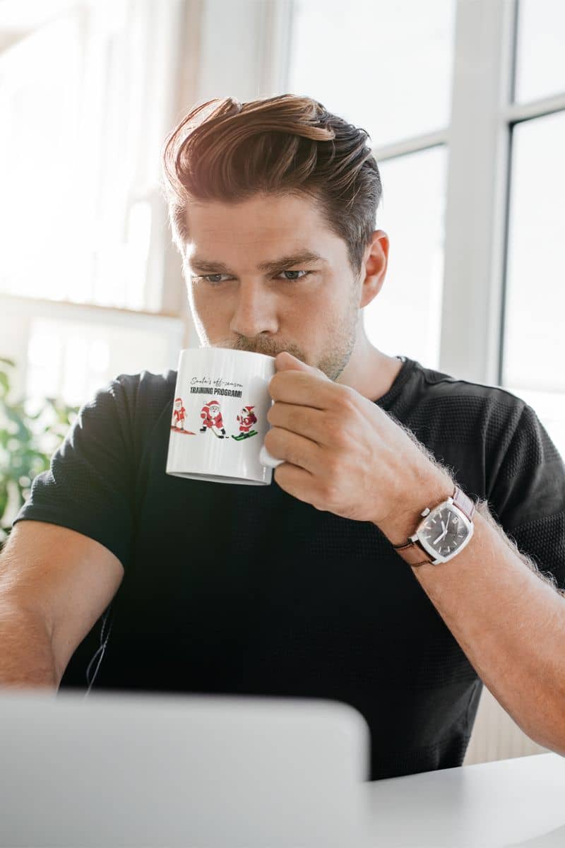 Santa's Off-Season Training Coffee Mug 11oz with a man drinking a cup of coffee while working on his laptop