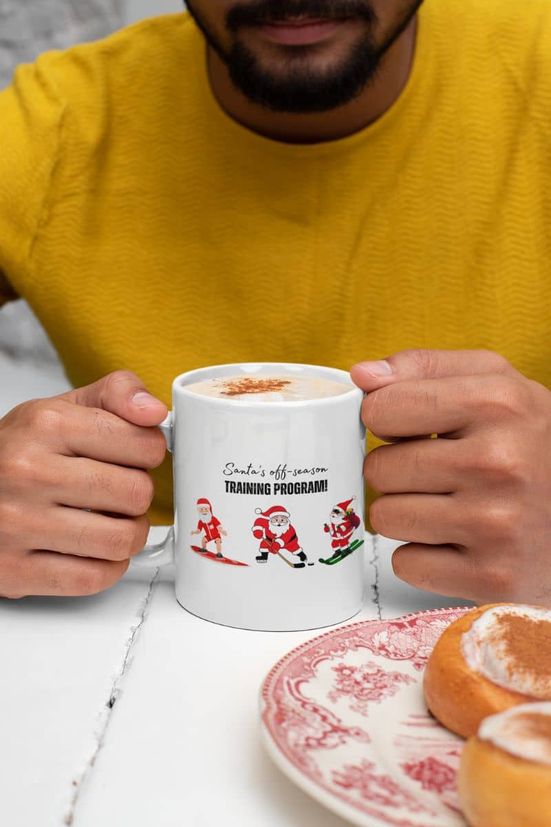 Santa's Off-Season Training Coffee Mug 11oz with a man holding a cup of coffee on the table next to a plate of cake