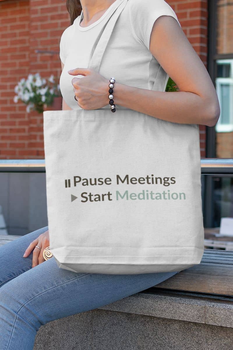 Sarcastic meditation tote with a person holding a tote bag with text 'Pause Meetings Start Meditation' on a blurred outdoor background