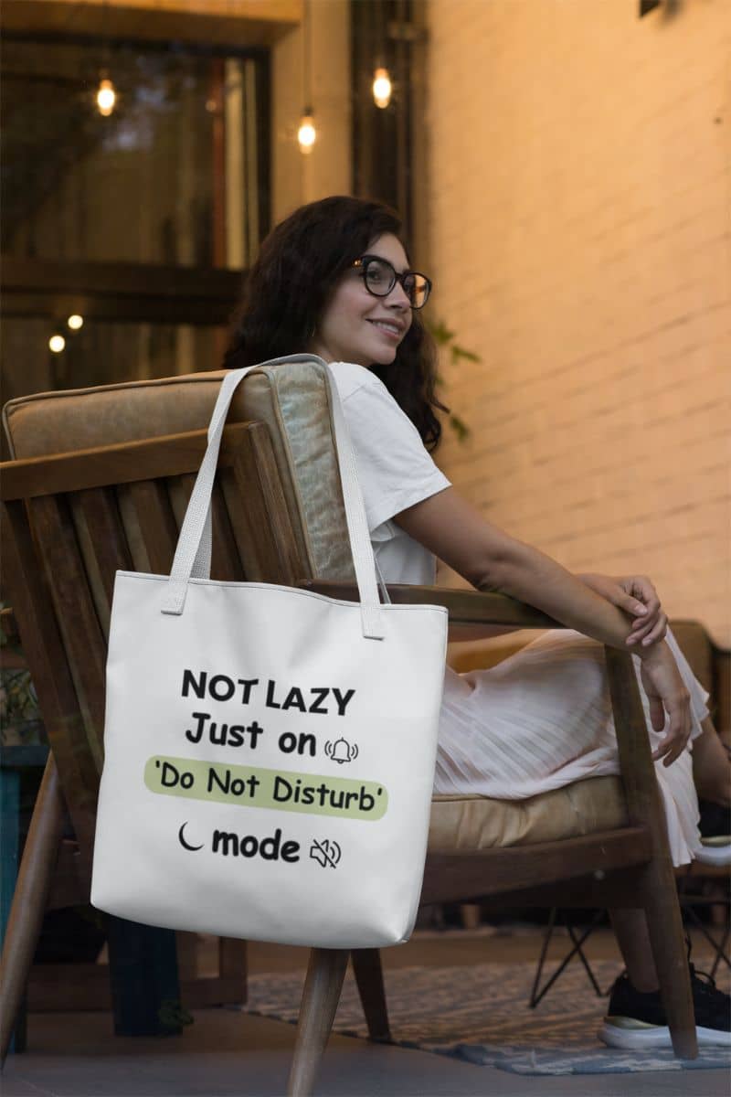 Sarcastic quiet mode tote with a woman sitting on a chair with a tote bag featuring humorous text 'NOT LAZY, Just on 'Do Not Disturb' mode'.