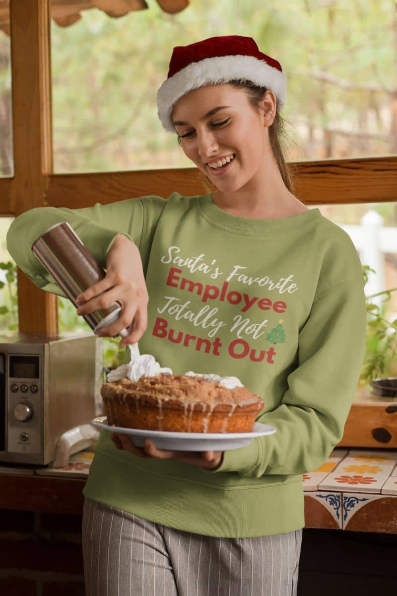 Sarcastic winter sweatshirt with a person in a green sweater with text 'Santa’s Favorite Employee Totally Not Burnt Out', holding a pie in a kitchen.