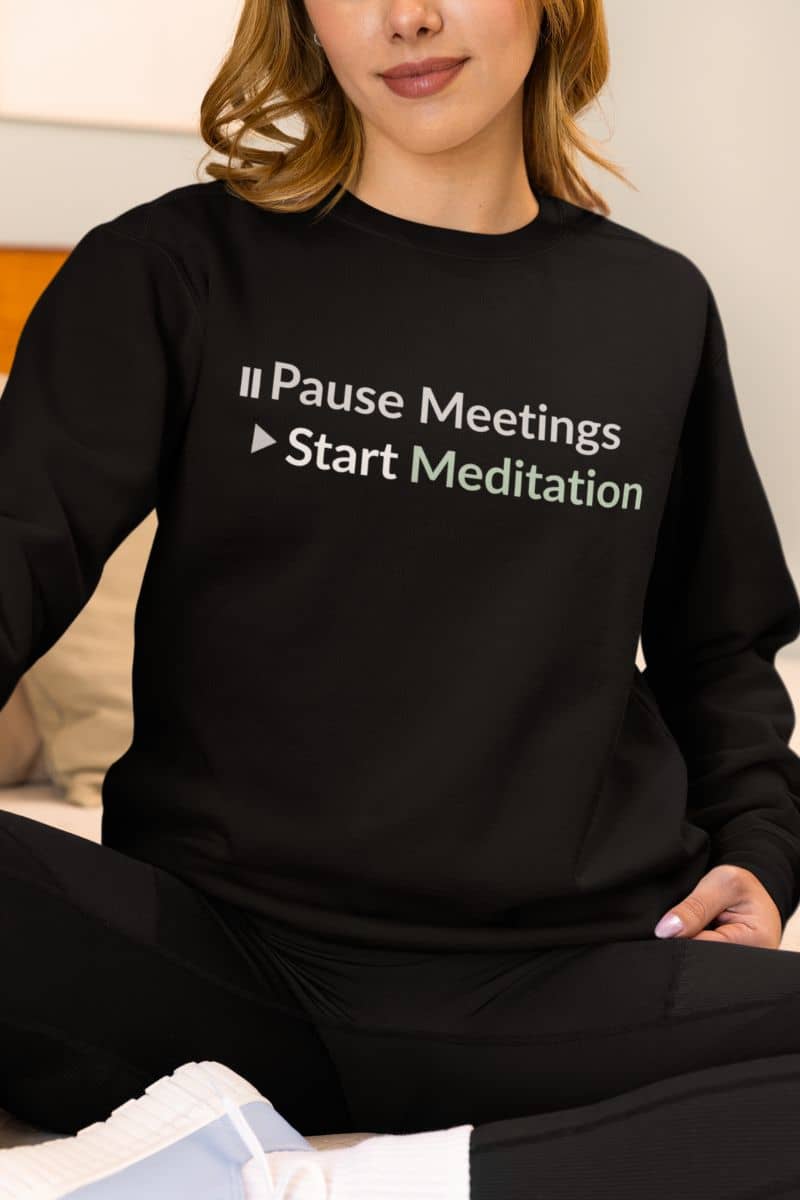Self-care motivational sweater with a person wearing a black sweatshirt with text 'Pause Meetings Start Meditation' sitting on a couch.