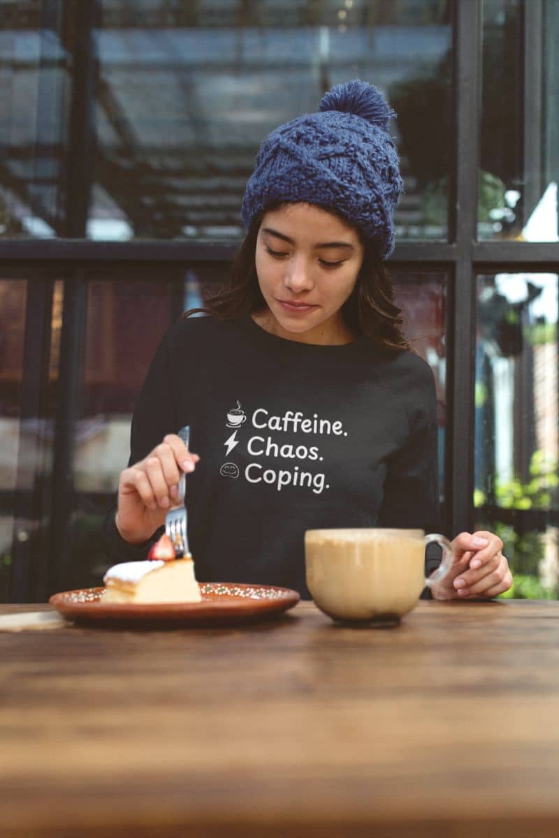 Self-care sweater with a person sitting at a table with a cup of coffee and dessert, wearing a black shirt with text 'Caffeine, Chaos & Coping'.