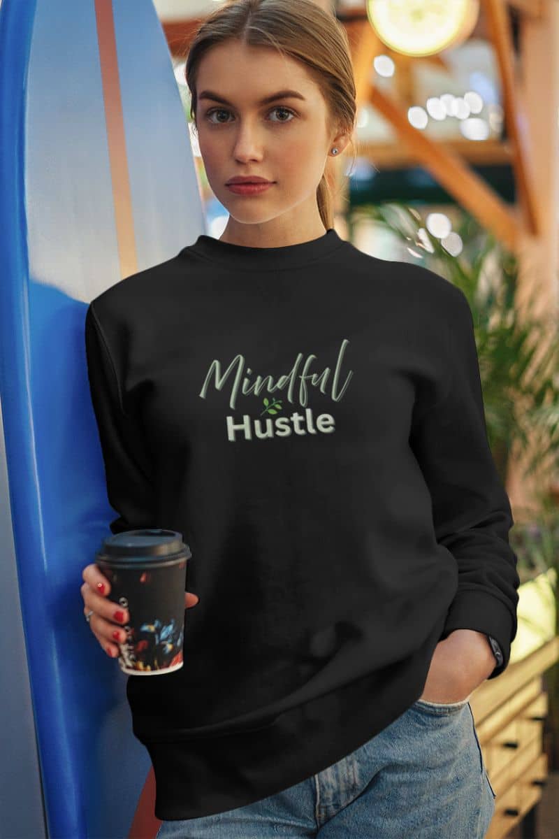 Self-development sweatshirt with a woman wearing a black sweatshirt with 'Mindful Hustle' text, holding a coffee cup, standing next to a blue surfboard.