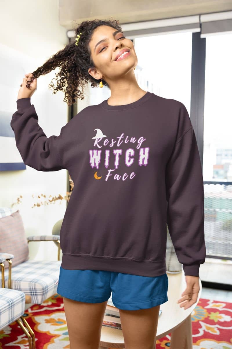 Spooky pullover hoodie with a woman displaying a sweatshirt with 'Resting Witch Face' text in a living room.