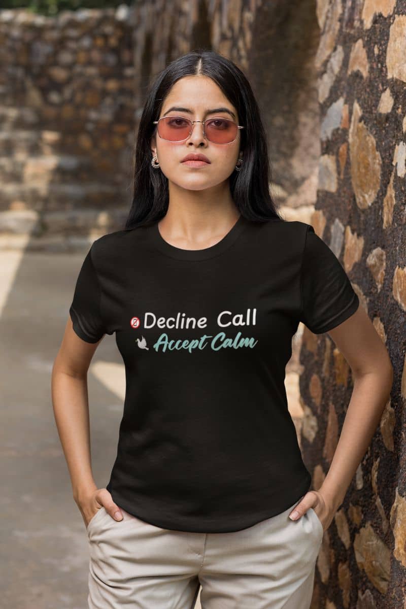 Stress relief t-shirt for women with a woman wearing a black t-shirt with text 'Decline Call Accept Calm', standing against a stone wall.