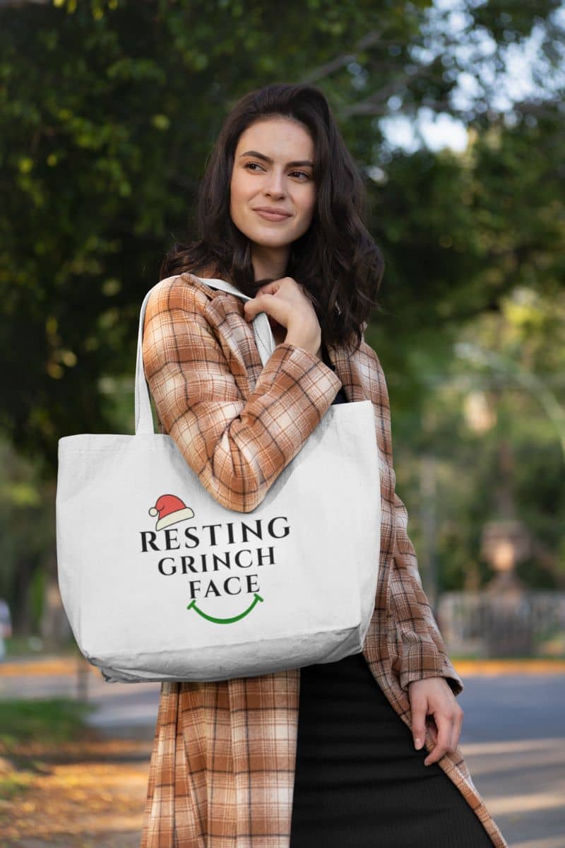 Subtle Christmas accessories with a woman holding a tote bag with 'Resting Grinch Face' text outdoors.