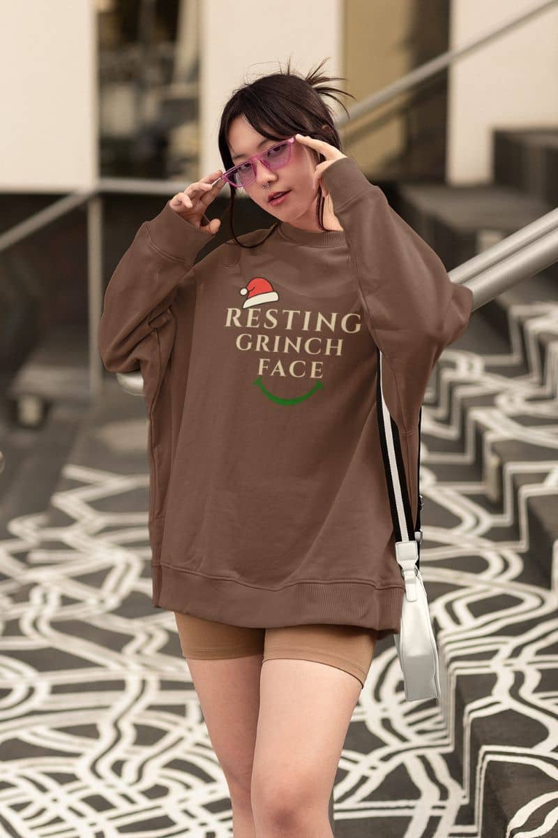 Winter sarcasm sweatshirt with a person wearing a brown sweatshirt with 'Resting Grinch Face' text, standing on a patterned floor.