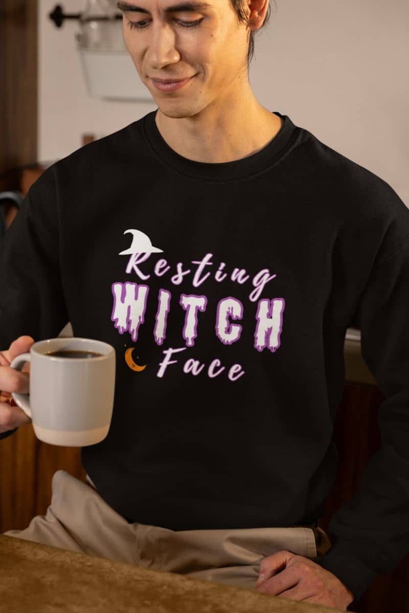 Witch humor pullover with a person wearing a black sweatshirt with 'Resting Witch Face' text, holding a mug.