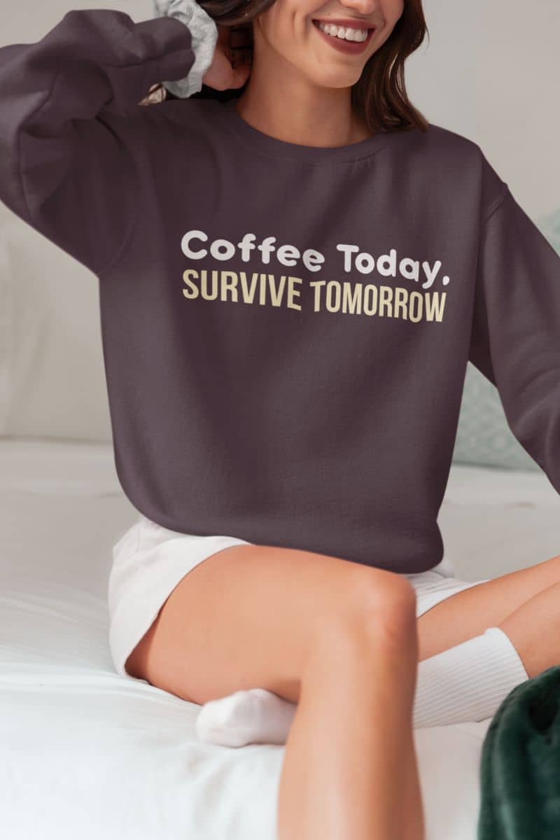 Workday motivation sweatshirt with a person in a brown sweatshirt with the text 'Coffee Today, Survive Tomorrow' on the bed.